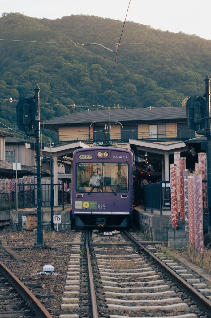 A purple Randen tram arriving at Arashiyama Station in Kyoto with wooded hills behind